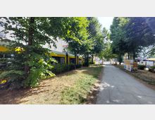 Schmaler Parkstreifen mit Fußweg, gesäumt von Bäumen und Sträuchern, daneben ein gelb-weißer Straßenbahnwagen. Im Hintergrund sind Häuser, parkende Autos und Werbeflächen sichtbar.