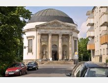 Klassizistisches Gebäude mit runder Kuppel und einem Säuleneingang, flankiert von zwei Skulpturen. Im Vordergrund eine Straße mit parkenden Autos, rechts angrenzende Wohngebäude und links von Bäumen gesäumt.