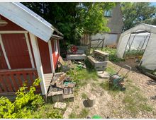 Ein Garten mit einer kleinen roten Holzhütte links, einer Werkbank und Gartengeräten in der Mitte sowie einem weißen Gewächshaus rechts. Im Hintergrund sind Bäume, Büsche und ein Teil eines grauen Gebäudes zu sehen.