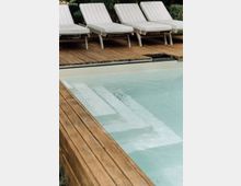 Ein Außenpool mit klarem Wasser und breiten weißen Stufen, umgeben von einem Holzdeck. Im Hintergrund stehen vier gepolsterte Liegestühle mit gestreiften Bezügen.