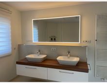 Modernes Badezimmer mit zwei rechteckigen Waschbecken auf einer hölzernen Arbeitsplatte, darunter weiße Schubladen. Ein großer, beleuchteter Spiegel hängt an der Wand, daneben ein weißer Handtuchheizkörper. Rechts und links sind weiße Wände mit schlichtem Dekor.