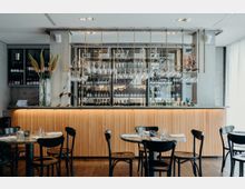 Moderne Bar in einem Restaurant mit hölzerner, vertikal geriffelter Thekenfront und einer Glas-Aufhängung für Weingläser. Im Hintergrund befinden sich Regale mit Flaschen, flankiert von Vasen mit dekorativen Pflanzen; davor stehen dunkle Stühle und Tische mit eingedecktem Geschirr.