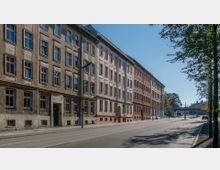 Straßenansicht mit einer Reihe von viergeschossigen Wohngebäuden im Altbau-Stil, linksseitig der Straße. Im Hintergrund ist eine Brücke sichtbar, und entlang der Straße stehen vereinzelt Bäume sowie Straßenlaternen.