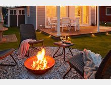 Gemütlicher Hinterhof mit einer kleinen Feuerstelle, umgeben von Kies, drei geflochtenen Stühlen mit Decken und einem kleinen Beistelltisch mit Getränken. Im Hintergrund ein beleuchtetes Holzdeck mit Esstisch und Stühlen neben einem hellgrauen Haus.