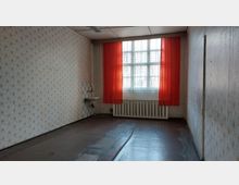 Ein leerer Raum mit vergilbter Tapete, einem Waschbecken in einer Ecke und einem großen Fenster mit roten Vorhängen. Unter dem Fenster befindet sich ein Heizkörper, und der Boden ist dunkel und abgenutzt.