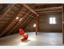 Dachboden mit sichtbaren Holzbalken, einem geneigten Ziegeldach und einem hellen Holzboden, beleuchtet durch ein kleines rechteckiges Fenster und ein halbrundes Fenster; in der Mitte steht ein einzelner orangefarbener Designerstuhl.