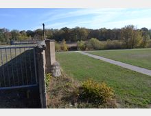 Blick auf eine Wiese mit einem Weg, der durch das Gelände führt, im Vordergrund ein altes Betonfundament mit einer Metallabsperrung. Im Hintergrund sind Bäume und Büsche in einer herbstlichen Landschaft zu sehen.