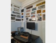 Ein Raum mit weiß gestrichenen Wänden, dekoriert mit einer Vielzahl von internationalen Kfz-Kennzeichen. An der Wand hängt ein Flachbildfernseher, darunter befinden sich ein Tisch mit einer schwarzen Verkleidung und diverse Batterien sowie technische Geräte.