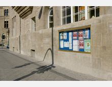 Fassade eines historischen Gebäudes aus beigem Stein mit hohen Fenstern und einem Aushangkasten mit Plakaten und Informationen. Der Gehweg aus grauen Steinplatten verläuft entlang der Wand, ein Laternenmast wirft einen Schatten auf die Fassade.