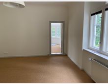 Leerer Raum mit beigefarbenem Teppichboden, weißen Wänden und einer Pendelleuchte an der Decke. Im Hintergrund befindet sich eine geöffnete Tür, die in einen weiteren, hell beleuchteten Raum mit großen Fenstern führt; rechts im Vordergrund sind ein Heizkörper und ein Fenster mit Blick ins Grüne sichtbar.