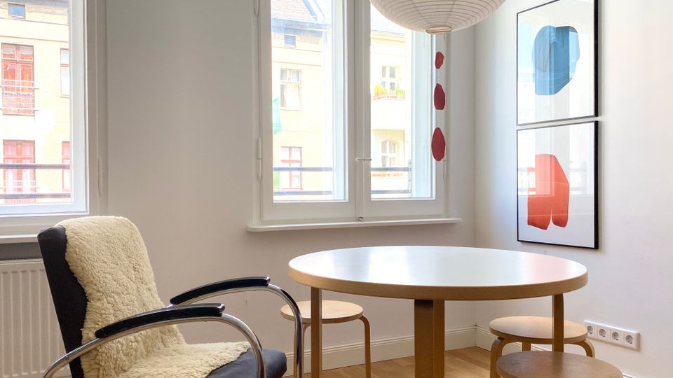 Heller Essbereich in einer Wohnung mit rundem Holztisch, kleinen Hockern und einem Sessel, daneben große hohe Fenster mit Blick auf gegenüberliegende Wohnhäuser. An der Wand hängen zwei gerahmte abstrakte Drucke, und von der Decke hängt eine große Papierlampion-Leuchte über dem Tisch.