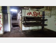 Ein verlassener, heruntergekommener Raum mit gefliesten Wänden, einem verrosteten Industriellen Ofen oder Maschinenblock im Vordergrund und Graffiti an den Oberflächen. Decke und Wände zeigen starke Spuren von Verfall und Verunreinigung.