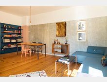 Wohnzimmer mit Holzfußboden, einer blauen Eckcouch und einem schwarzen Couchtisch. Im Hintergrund befinden sich ein Esstisch mit Stühlen, eine blaue Bücherwand sowie eine kleine Holzkommode; die Wände sind in Betonoptik mit moderner Kunst dekoriert.