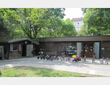 Holzgebäude in einem Park mit Bäumen im Hintergrund. Vor dem Gebäude stehen Kinderfahrzeuge wie Go-Karts und Fahrräder sowie ein Regal mit bunten Helmen.