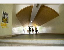 Ein überdachter Fußgängertunnel mit gelblichen Ziegeln an der gewölbten Decke und hell gefliesten Wänden. Im Tunnel laufen mehrere Personen in Richtung eines hell beleuchteten Ausgangs; an der Wand ist ein beschmiertes Plakat zu sehen.