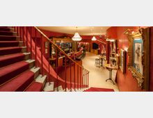 Ein stilvoll eingerichteter Barbereich in einem Theater oder Varieté mit roten Wänden, verzierten Gemälderahmen, einer geschwungenen Treppe mit rotem Teppich und eleganten Kronleuchtern. Im Hintergrund befinden sich eine lange Bar, hohe Hocker und runde Tische mit Barhockern.