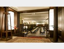 Innenraum eines traditionellen Restaurants mit dunklen Holzsäulen, gedeckten Tischen und einem Teppich im vorderen Bereich. Große Lampen an den Wänden und eine leicht erhöhte Plattform führen in den Speisesaal.