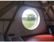 Ein Innenraum mit sichtbaren Holzbalken, einem roten Fußboden und einem großen, runden Fenster, das den Blick auf eine grüne Wiese mit Bäumen freigibt. Im Vordergrund stehen ein graues Sofa und ein blauer Sessel.