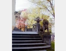 Steintreppe mit schmiedeeisernen Geländern, flankiert von dekorativen Steinvasen und einer Löwenskulptur, vor einem Hintergrund aus blühenden Bäumen und einem angrenzenden Gebäude.