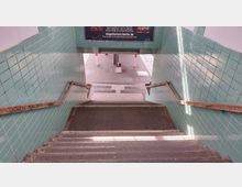 Treppenabgang in eine U-Bahn-Station mit Wänden aus grünlichen Fliesen, Holzhandläufen und einem Plakat mit Werbung im oberen Bereich. Am Ende der Treppe führt der Zugang zu einem gepflasterten Platz.