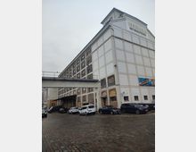 Historisches Speichergebäude mit der Aufschrift "Behala Viktoriaspeicher", bestehend aus Mauerwerk und großen weißen Fassadenelementen; im Vordergrund eine gepflasterte Fläche mit geparkten Autos und ein erhöhter Verbindungsgang.