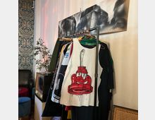 Ein Innenraum mit einer Kleiderstange voller bunter Kleidungsstücke, darunter ein auffälliges weißes Tanktop mit einer roten Comic-Figur. Im Hintergrund befinden sich eine gemusterte Tapete, ein kleiner Fernseher in Retro-Design, eine Pflanze und ein Schwarz-Weiß-Bild an der Wand.