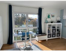 Helles Kinderzimmer mit einem großen Fenster, blauen Vorhängen und Blick auf eine grüne Landschaft. Im Raum stehen ein weißer Schreibtisch mit einem blauen Bürostuhl, ein Regal mit Spielzeug und Aufbewahrungsboxen sowie eine elektrische Tastatur auf einem Rollwagen.
