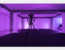 Ein Studio mit schwarzem Bodenbelag und einem dreibeinigen Kamerastativ im Vordergrund. Der Raum ist mit Metallgerüsten und mehreren kleinen Leuchten ausgestattet, in violettem Licht beleuchtet.