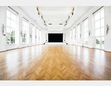 Großer, heller Veranstaltungssaal mit hellem Parkettboden, hohen weißen Wänden und großen Fenstern, die viel Tageslicht hereinlassen. Am Ende des Saals befindet sich eine Bühne mit schwarzem Vorhang, flankiert von dekorativen Wandleuchten.
