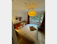 Schlafzimmer mit einem Doppelbett aus Rattan, dekoriert mit bunten Kissen und einer weißen Tagesdecke. Über dem Bett hängt eine Wanddekoration, und ein Fenster mit roten Vorhängen bietet Ausblick auf einen Garten mit Steinen und Grünpflanzen; eine große, gelbe Hängeleuchte dominiert die Decke.