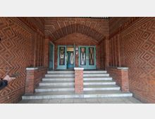 Überdachter Eingangsbereich eines Backsteingebäudes mit gemustertem Ziegelgewölbe, breiter Treppe und mittigem Ziegelpfeiler. Oben befinden sich grün gestrichene Doppeltüren mit verglasten Einsätzen und Rautengittern.