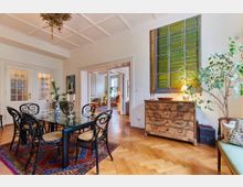 Esszimmer mit einem Glastisch und sechs schwarzen Stühlen auf einem roten Teppich, einem Holzboden und einer antiken Kommode. Im Hintergrund sind eine geöffnete Tür zu einem hellen Wohnzimmer sowie große Fenster erkennbar.