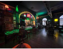 Innenraum einer irisch gestalteten Bar mit dunklen Wänden, hohen Tischen und Barhockern sowie Holzfässern als Stehtische. Im Hintergrund sind eine kleine Bühne mit beleuchtetem Torbogen, Leuchtreklamen und große Rundbogenfenster zu sehen.