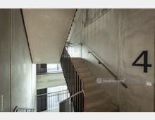 Betontreppe in einem modernen Treppenhaus mit schlichten Wänden und metallenen Handläufen, beleuchtet durch Tageslicht von einem Fenster. An der Wand ist groß die Zahl "4" angebracht.