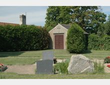 Kleine Friedhofskapelle mit braunen Doppeltüren und einem Kreuz an der Fassade, umgeben von Bäumen, Hecken und einer Rasenfläche; im Vordergrund sind ein Grabstein, ein großer Stein und blühende Grabbepflanzungen sichtbar.