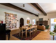 Wohnzimmer mit Holzboden, Holzbalkendecke und einem großen Esstisch in der Mitte, umgeben von Holzstühlen. An der linken Wand befindet sich ein Bücherregal, rechts ein gemauerter Kamin mit braunen Ziegeln, kombiniert mit einem Ledersofa und einem Sessel. Ein Flügel steht in der linken Ecke, und der Raum ist durch eine große Glastür zum Außenbereich hin offen.