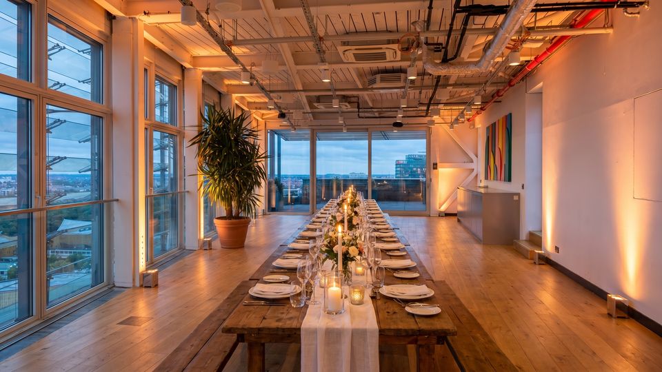 Modern loft-style event space with floor-to-ceiling windows overlooking a city, wood floors, and an exposed industrial ceiling with ducts and lighting. A long banquet table is set with plates, glassware, candles, and floral arrangements, with a large potted plant and wall art along the side.