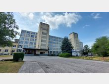 Großes beige-farbenes Verwaltungs- oder Industriegebäude im funktionalistischen Stil mit hohen, zentralen Türmen und vielen rechteckigen Fenstern. Im Vordergrund ein gepflasterter Zufahrtsweg, grüne Büsche, ein Nadelbaum und Hinweisschilder; umgeben von Wiesen und Bäumen.