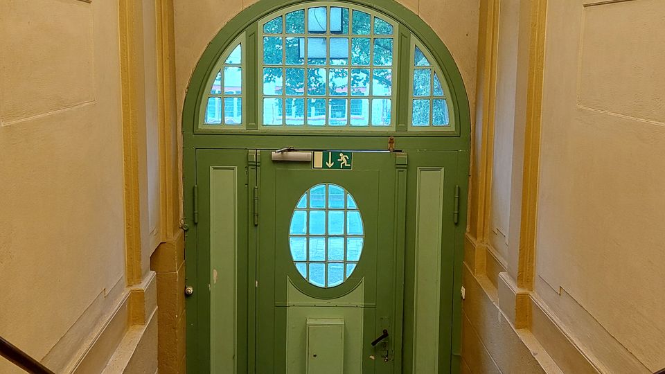 Treppenhaus mit einer grünen Holztür und ovalem Fenster im Eingangsbereich. Über der Tür befindet sich ein buntes Glasfenster mit abstraktem Motiv, während die Raumdecke mit einem weißen Gewölbe und vielen kleinen Leuchten verziert ist.