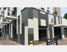 Mehrstöckiges modernes Gebäude mit einer Fassade aus vertikal angeordneten grauen und weißen Paneelen. Im Vordergrund befinden sich gläserne Eingangstüren, schmale Balkone mit Metallgeländern und ein Schild mit der Aufschrift „Allee der Kosmonauten 25A“.