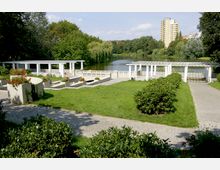 Parklandschaft mit einem gepflegten Rasen, Blumenbeeten und weißen Pavillons mit Pergolen. Im Hintergrund ein ruhiger See, umgeben von Bäumen, sowie einige mehrstöckige Gebäude.
