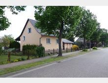 Beige Einfamilienhaus mit grauem Satteldach, umgeben von einem Garten mit Hecken und Blumen, entlang einer von Bäumen gesäumten Straße mit Bürgersteig und schmiedeeisernem Zaun. Im Hintergrund sind Nachbarhäuser und weitere Vegetation sichtbar.