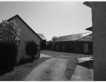 Schwarz-weiß-Aufnahme eines ländlichen Hofs mit mehreren Gebäuden. Links ist ein kleines Haus mit Satteldach und Büschen davor, rechts ein größeres Backsteingebäude mit einfachem Dach; dazwischen ein unbefestigter Weg und eine Grasfläche.