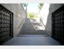 Breiter Treppenaufgang aus Beton mit Geländern auf beiden Seiten, führt aus einer Unterführung zu Tageslicht. Links und rechts befinden sich verzierte Metalltüren, und oben ist ein Schild mit dem U-Bahn-"U"-Symbol sichtbar.