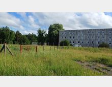 Eine Wiese mit hohem Gras, im Hintergrund ein schmuckloses, graues, mehrstöckiges Gebäude mit regelmäßigen Fensterreihen. Links und im hinteren Bereich sind Zäune sowie vereinzelte Bäume zu sehen, die an eine bewaldete Umgebung grenzen.