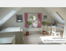 Helles Dachzimmer mit einer schrägen Decke, einem runden Fenster mit rosa Vorhängen, einem weißen Bett links und einem Schreibtisch mit Stuhl rechts. Zwei Topfpflanzen stehen vor dem Fenster, und helle Holzböden dominieren den Raum.