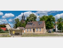 Straßenszene mit einem kleinen Backsteinhaus mit Satteldach und Solarpanels, umgeben von Bäumen und Nachbarhäusern. Im Hintergrund ragt ein Kirchturm mit einer schwarzen Spitze auf, im Vordergrund ist eine einfache Bushaltestelle mit einer Bank zu sehen.