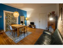 Wohnzimmer mit Holzboden, blauer Wand und hölzernem Esstisch, umgeben von verschiedenen Stühlen. Rechts stehen ein brauner Kachelofen, eine schwarze Ledercouch und ein TV auf einem Holzregal; eine helle Stehlampe und ein großer Spiegel ergänzen den Raum.