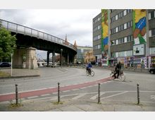 Straßenszene in einer Stadt mit einem erhöhten Bahngleis auf massiven Steinpfeilern links und einem großen Gebäude rechts, das mit Wandgemälden geschmückt ist. Im Vordergrund fahren zwei Personen auf Fahrrädern einen Radweg entlang, und im Hintergrund sind die Türme der Oberbaumbrücke sichtbar.