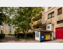 Mehrfamilienhaus mit gelber und beige gestrichener Fassade, Balkonen und einem überdachten Bereich für Mülltonnen. Im Vordergrund stehen Bäume und eine gepflasterte Gehwegfläche, im Hintergrund sind weitere Wohnblöcke sichtbar.